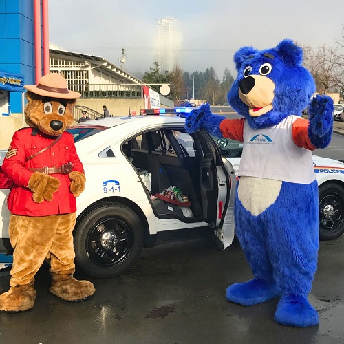  RCMP Safety Bear standing with their arms in the air on one side of a marked RCMP police car with the SHARE Society Bear standing on the other side with their arms in the air