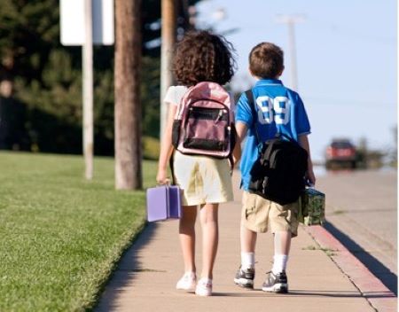 An image taken from behind of a boy and a girl walking on a sidewalk wearing backpacks.  