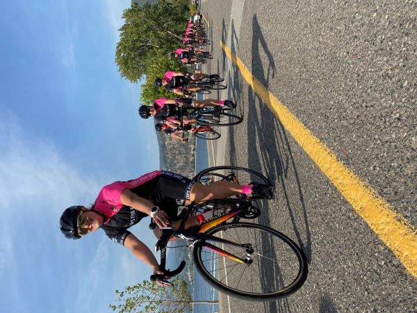 Un groupe de cyclistes vêtus d’un uniforme noir et rose roule sur une route avec des montagnes en arrière-plan.