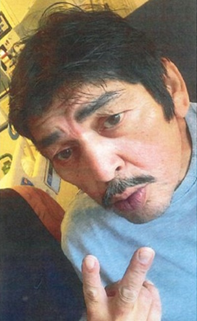 Photo of Alfred Dumais with black hair and a greying mustache, wearing a grey crew neck shirt, giving a peace sign