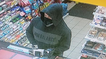 A Caucasian man in a black sweater with New Balance written on it and a black mask is front of a counter at a convenience store. 