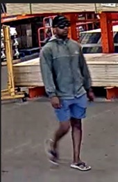 surveillance photo of suspect wearing a black baseball hat, sunglasses, long-sleeve grey zippered jacket and blue shorts.