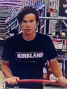 male suspect pushing a cart inside the store. He is wearing a black t-shirt with <q>Kirkland</q> in white writing on the front. He has short brown hair swooped to the side.