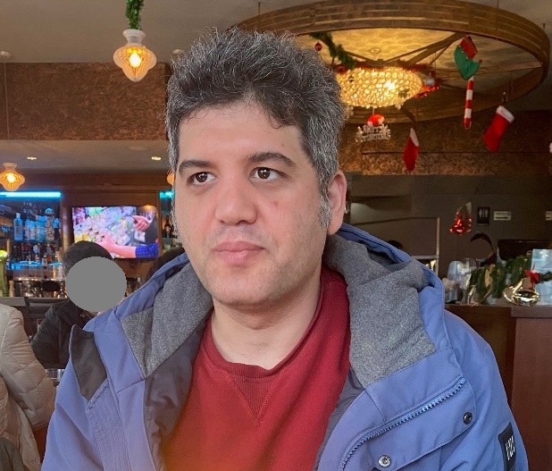 Middle Eastern man with greying hair and brown eyes, wearing a blue winter jacket and a red shirt.