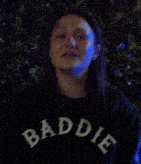 A blurry image of a Caucasian female with brown hair and brown eyes, wearing a dark sweater with the word <q>BADDIE</q> written across the chest.