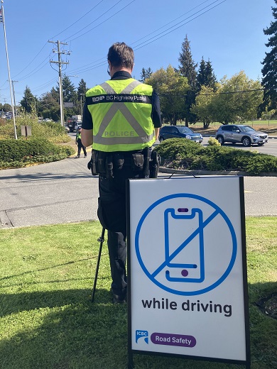 Same rear view of previous officer, with an ICBC/RoadSafetyBC sign in foreground displaying a picture of a crossed-out cellphone